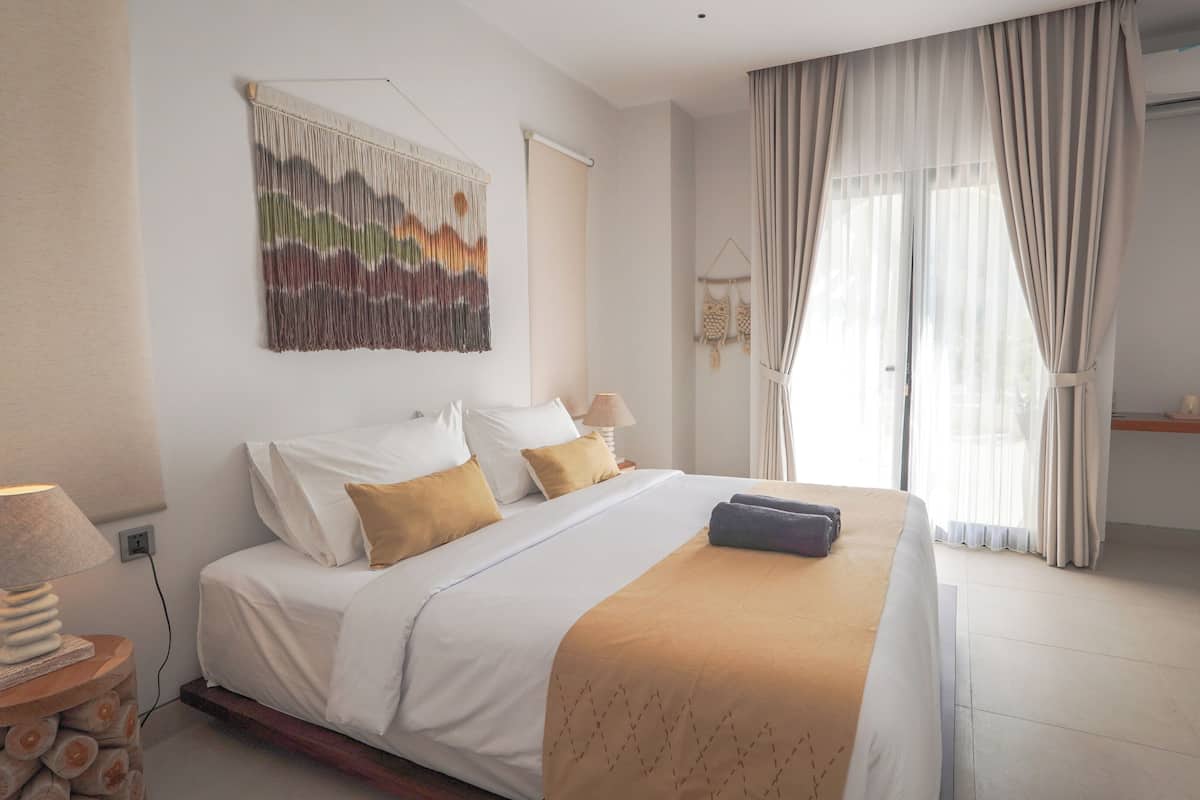Quarto de hotel com cama, obra de arte na parede, mesa de cabeceira com abajur e janela com cortinas.
