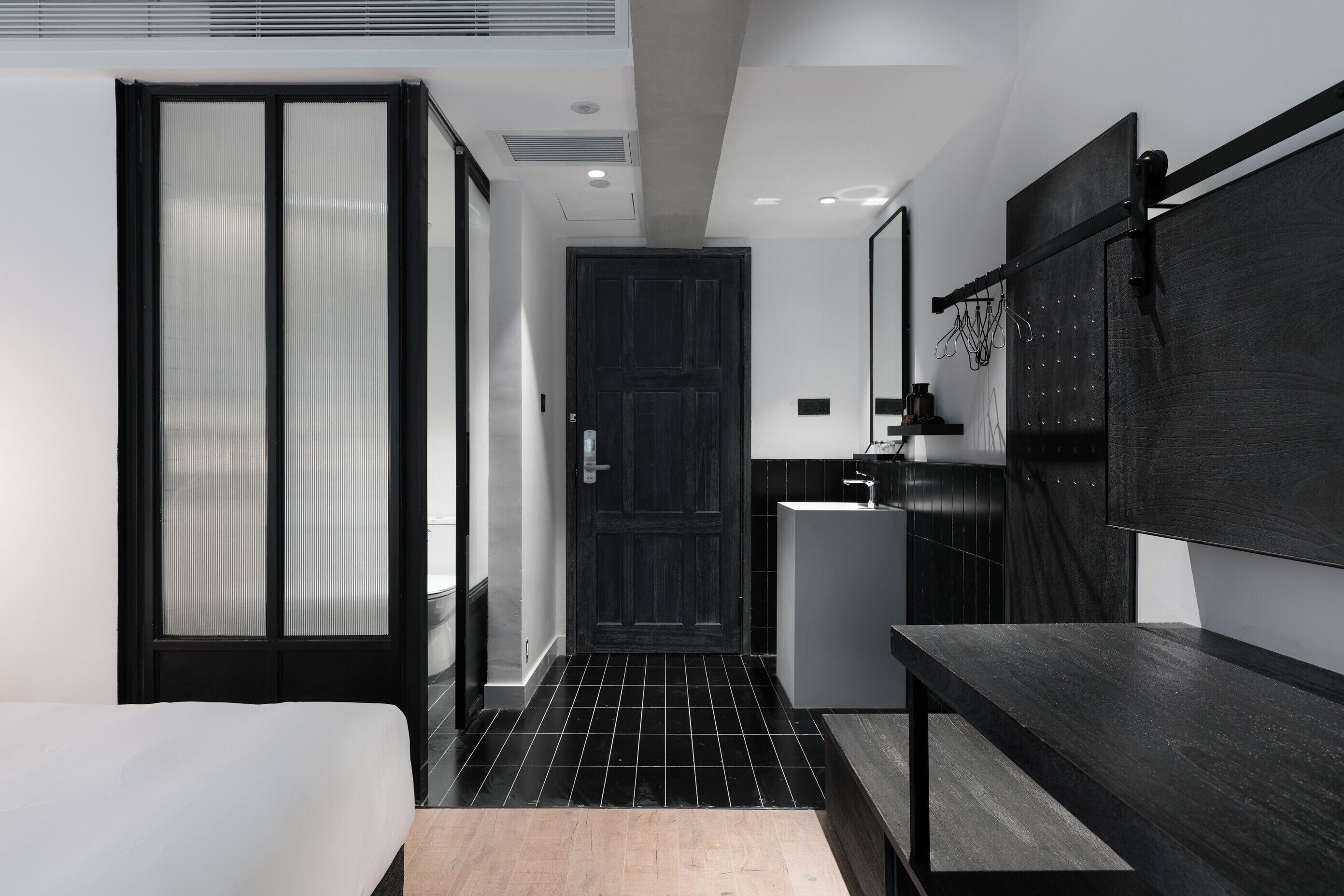 A modern hotel room with a bed, a wooden bench, a black-framed door, a small sink, and a TV mounted on the wall.
