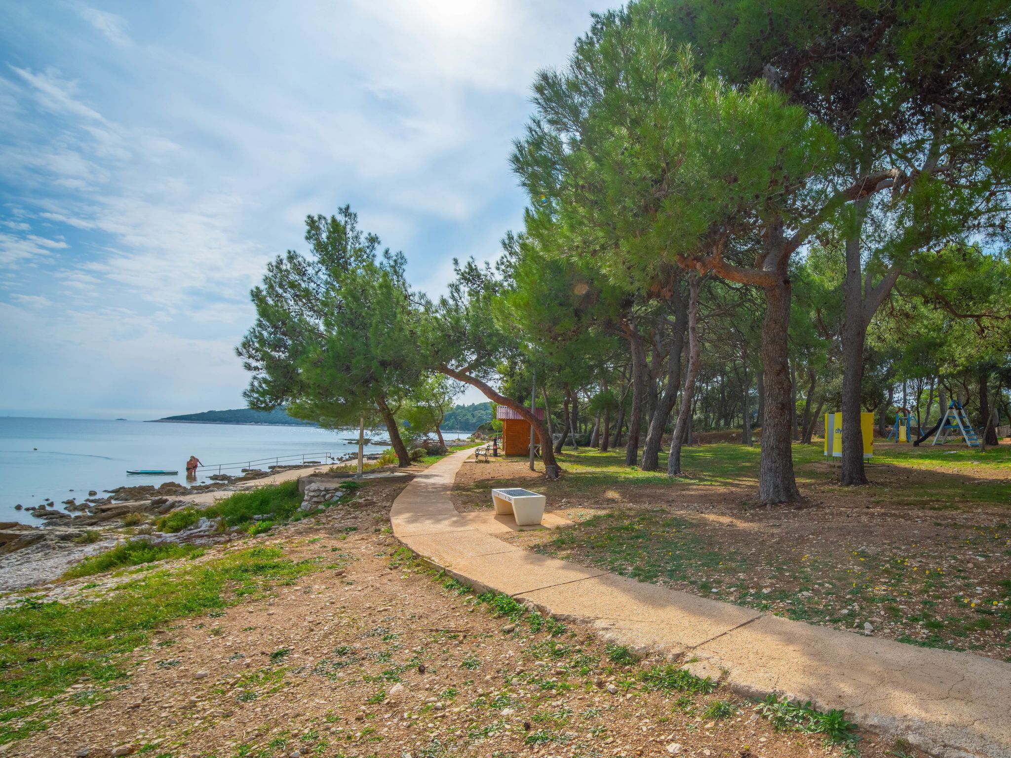 A coastal park with a walking path, trees, and a small wooden structure.