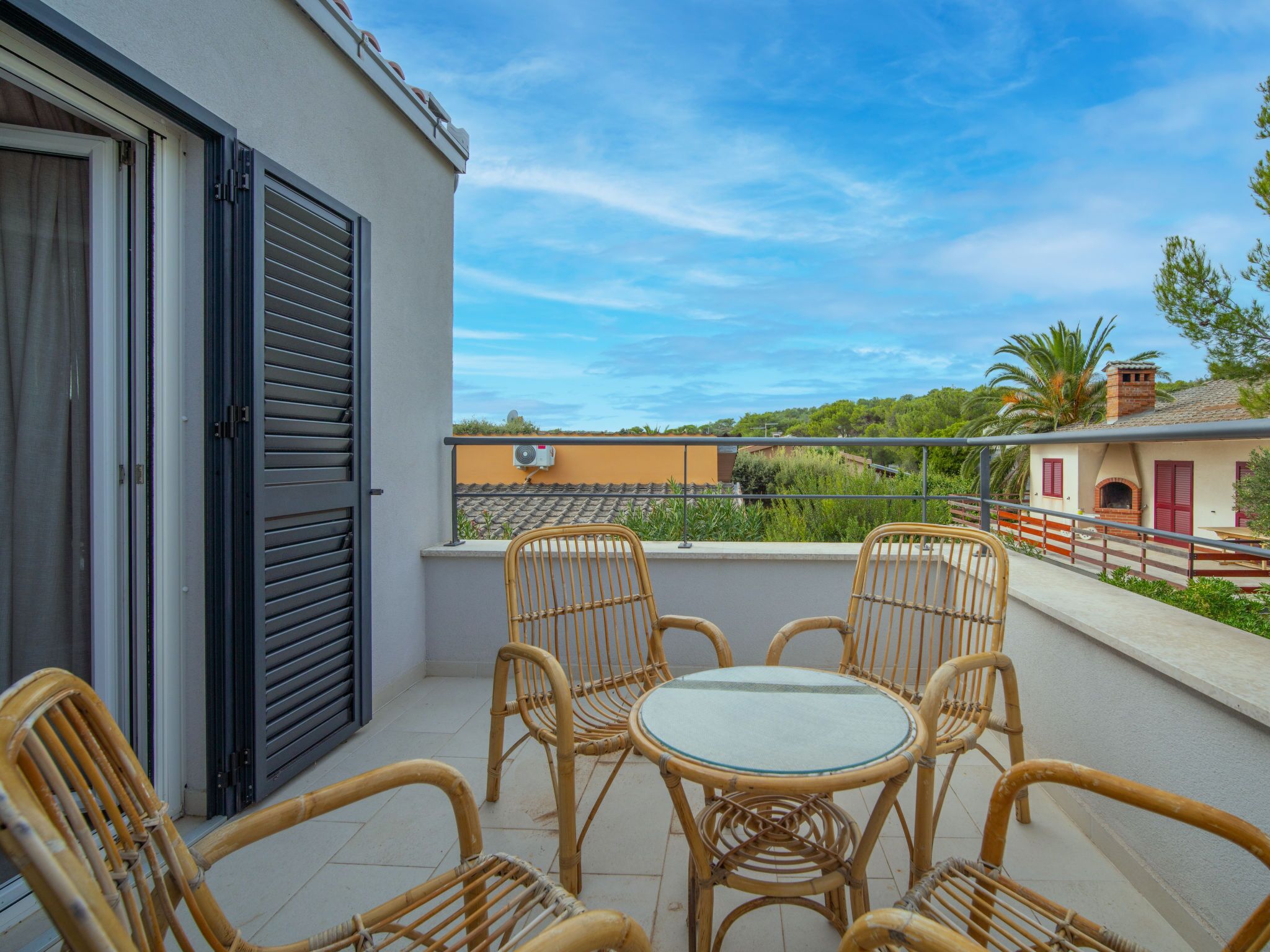 A balcony with wicker chairs and a table, overlooking a residential area with houses and greenery.
