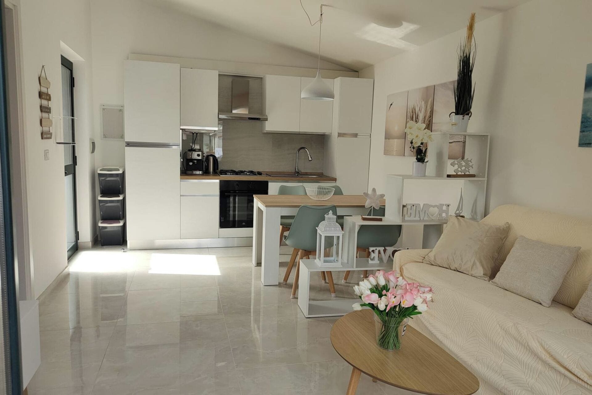 A modern living room with a beige sofa, a wooden coffee table with flowers, and a kitchen area with white cabinets and a dining table.