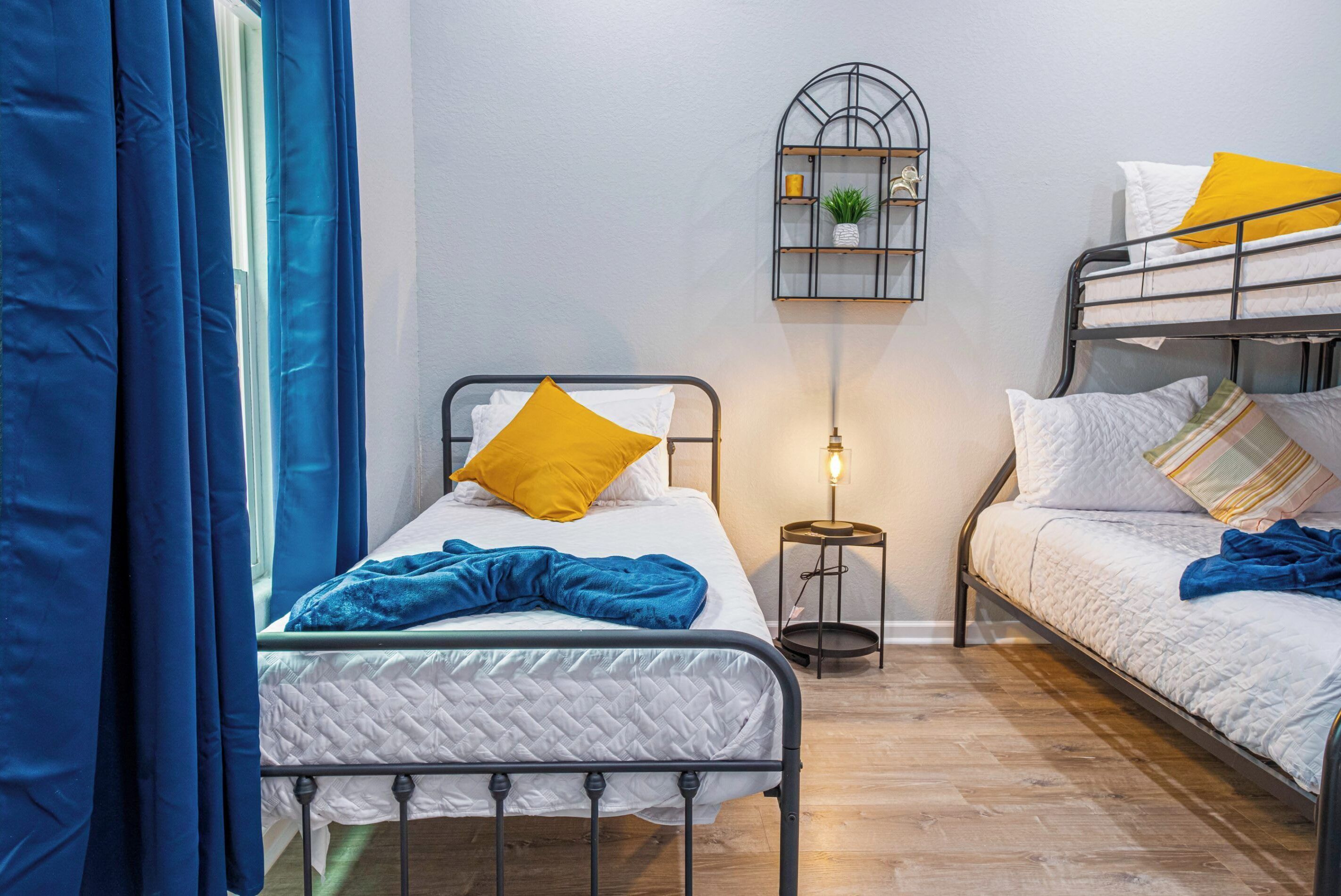 A dormitory room with two bunk beds, a bedside table with a lamp, and a wall-mounted shelf with decorative items.