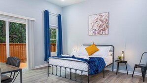 Un dormitorio con cama, escritorio, silla, una mesita con lámpara y ventana con cortinas.