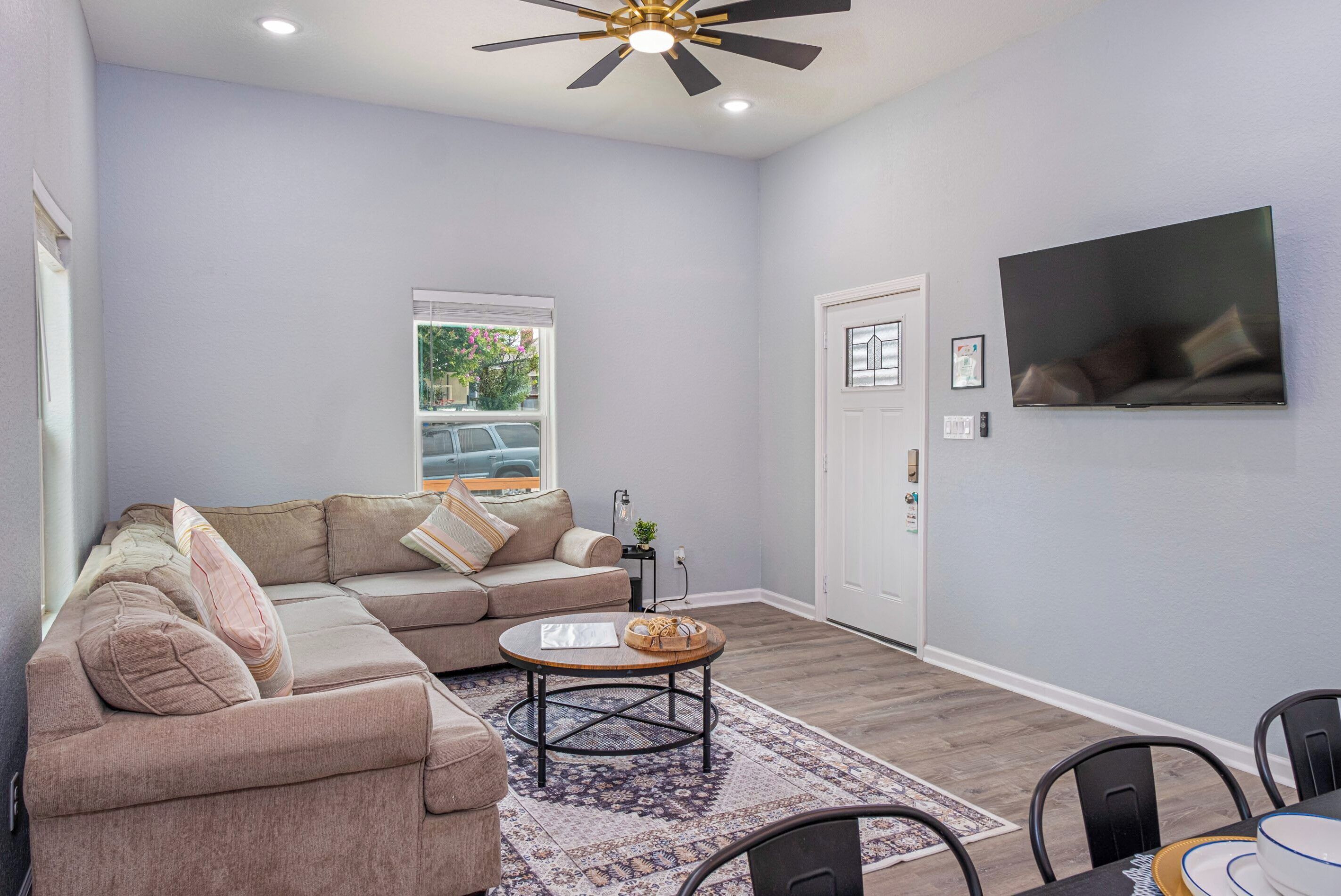 A living room with a sectional sofa, a coffee table, a TV mounted on the wall, and a ceiling fan.