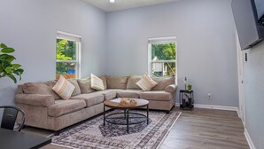Una sala de estar con un sofá modular, una mesa de centro redonda y un ventilador de techo. Hay dos ventanas con persianas, un televisor montado en la pared y una mesita auxiliar con una planta.