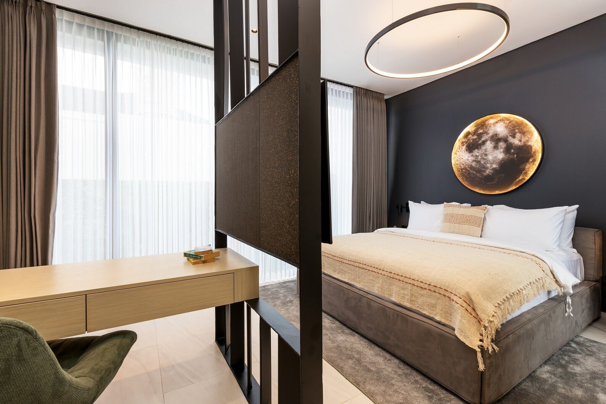 Une chambre d’hôtel moderne, dotée d’un bureau, d’un lit recouvert d’une couverture beige et d’un grand élément décoratif circulaire au mur.