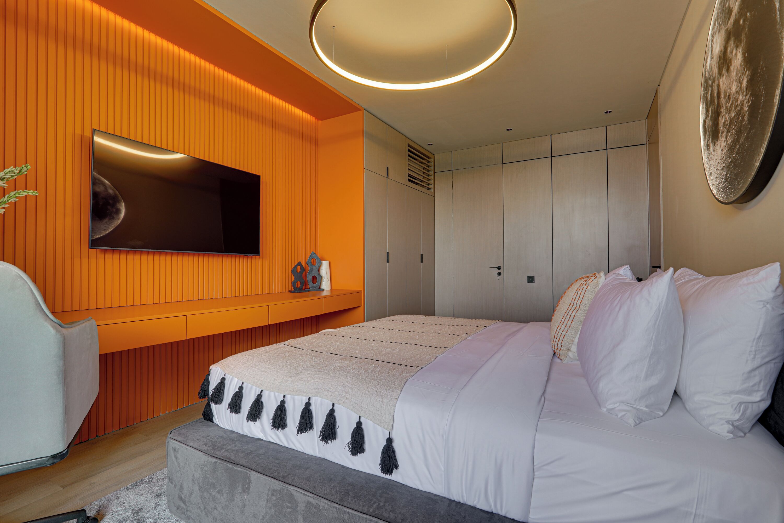 Ein modernes Schlafzimmer mit einem großen Bett, einem an einer orangefarbenen Wand montierten Fernseher und einem Kleiderschrank mit Schiebetüren.