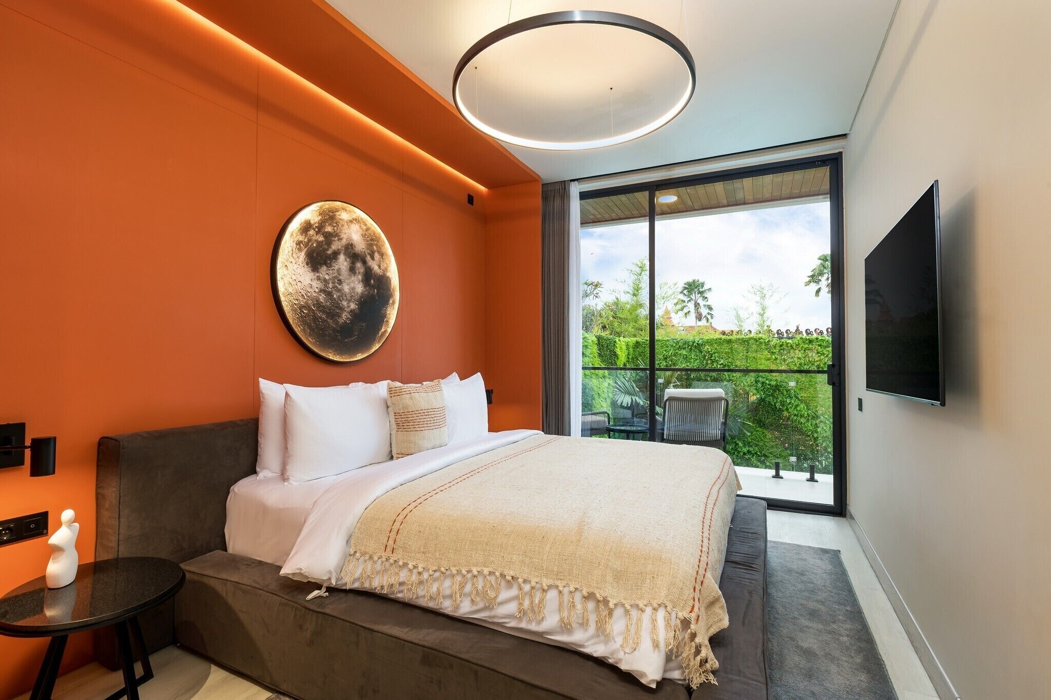 A modern bedroom with a large bed, a round wall art piece, a TV mounted on the wall, and a balcony with a view of greenery.