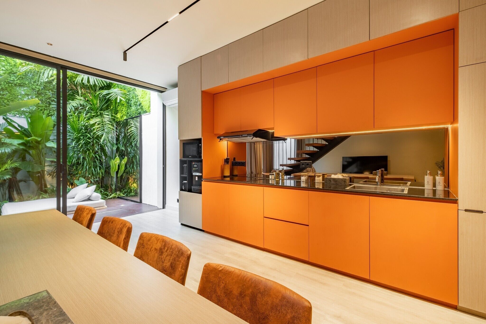 A modern kitchen with orange cabinetry, a dining area with a wooden table and chairs, and a view of lush greenery outside.
