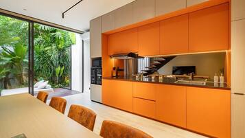 A modern kitchen with orange cabinetry, a dining area with a wooden table and chairs, and a view of lush greenery outside.
