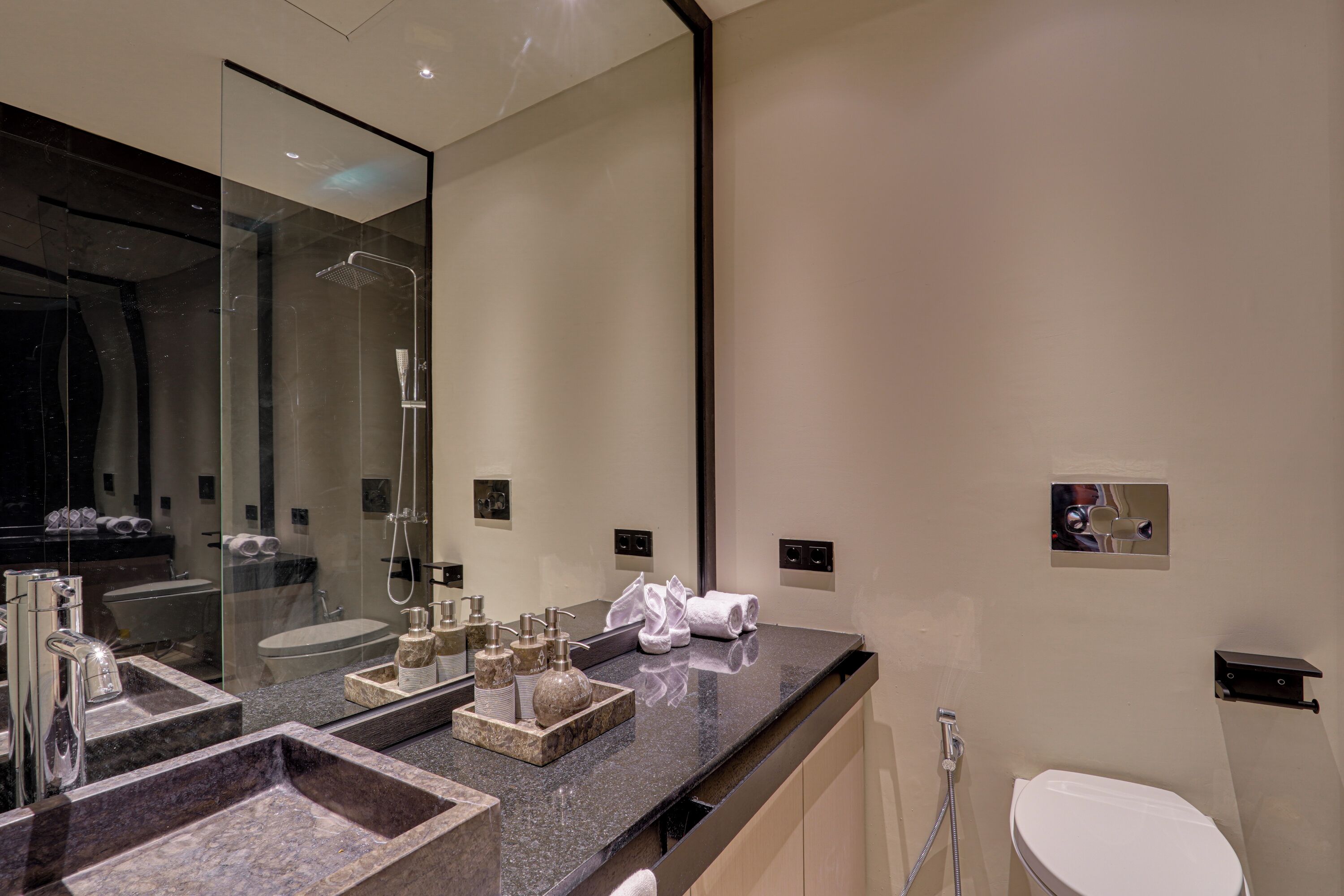 Un baño moderno con un espejo grande, ducha con cerramiento de vidrio, lavamanos con varios recipientes y un inodoro.