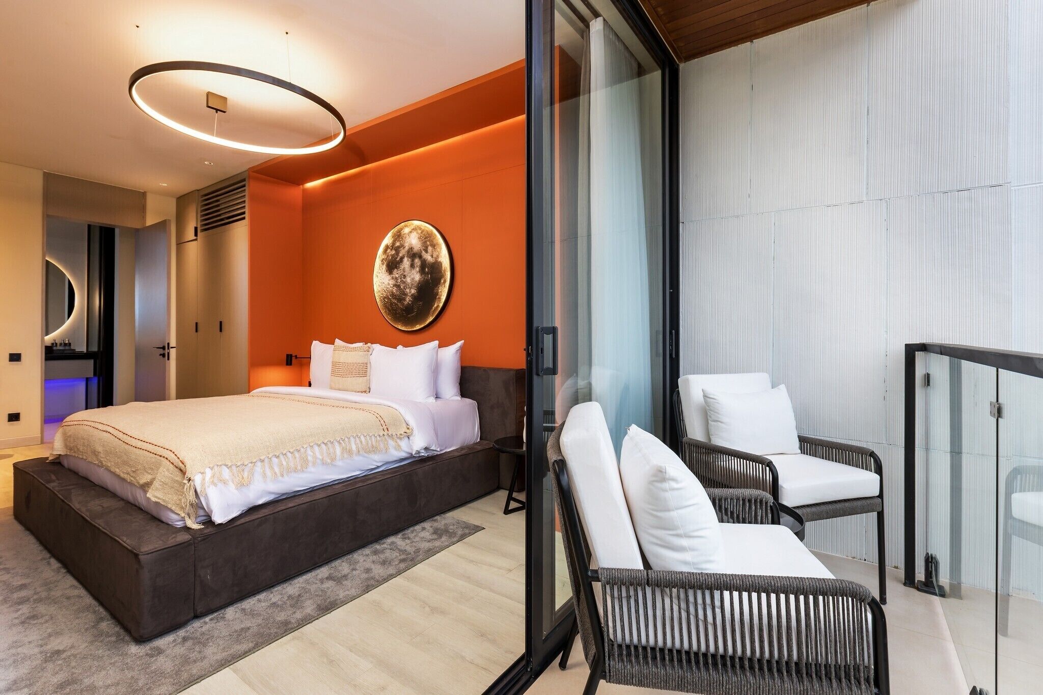 A modern bedroom with a large bed, a circular light fixture, and a glass partition separating the room from a balcony area with wicker furniture.