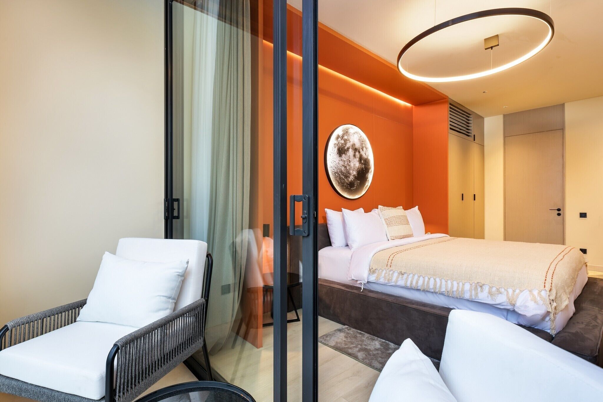 Une chambre d’hôtel moderne avec une cloison en verre, un lit avec une literie blanche, un fauteuil en osier et un luminaire circulaire.