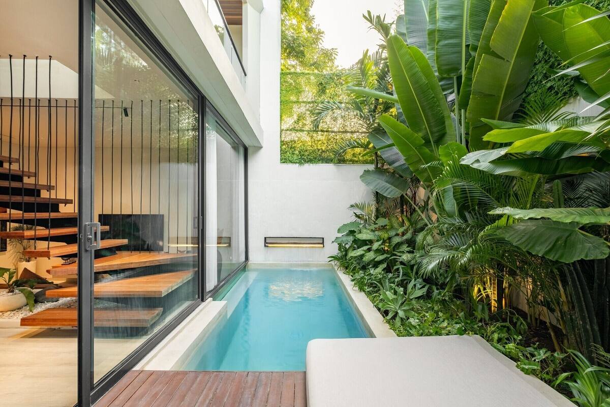 Un área de piscina moderna al aire libre con cubierta de madera, paredes de vidrio y exuberante vegetación tropical.
