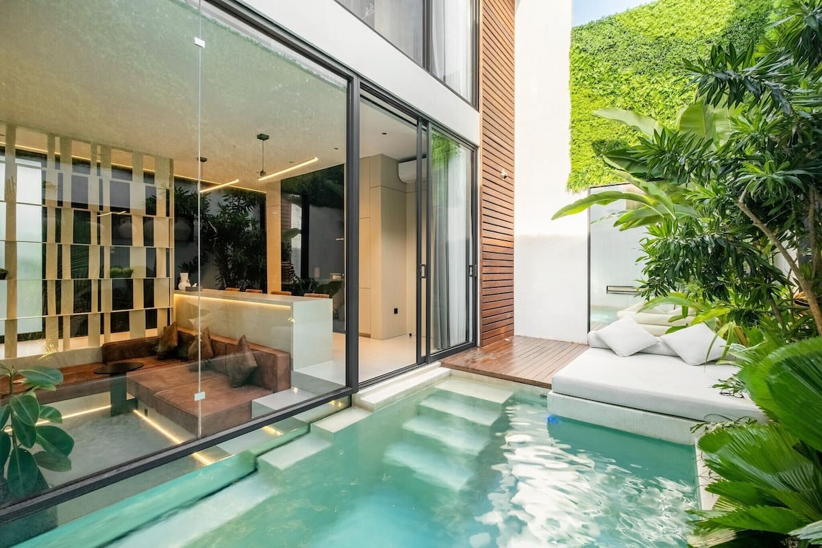 Una casa moderna con un salón envuelto en vidrio, una piscina y abundante vegetación.
