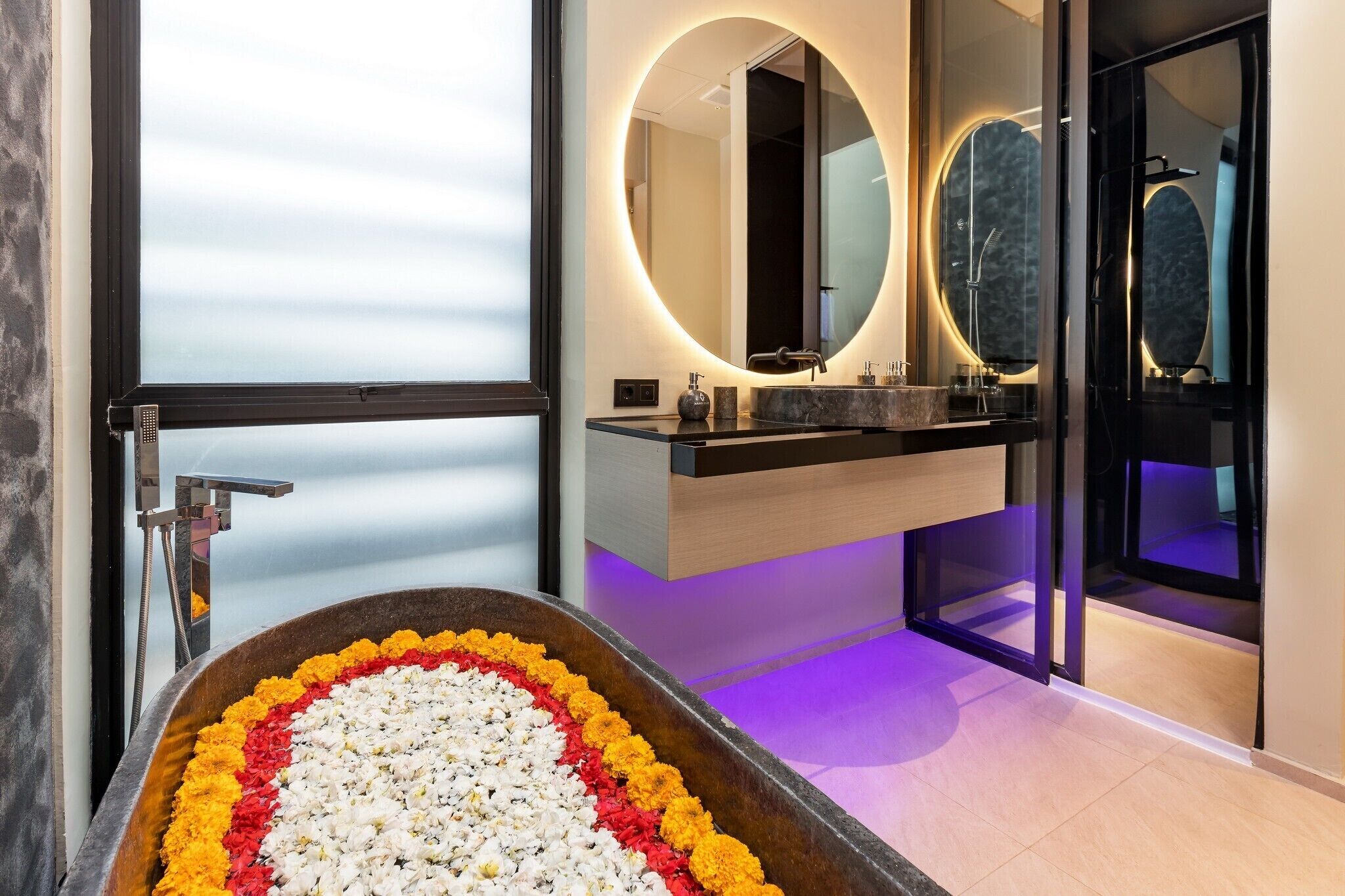 Un baño moderno con un espejo ovalado grande, una bañera con pétalos de flores y un box de ducha de vidrio.