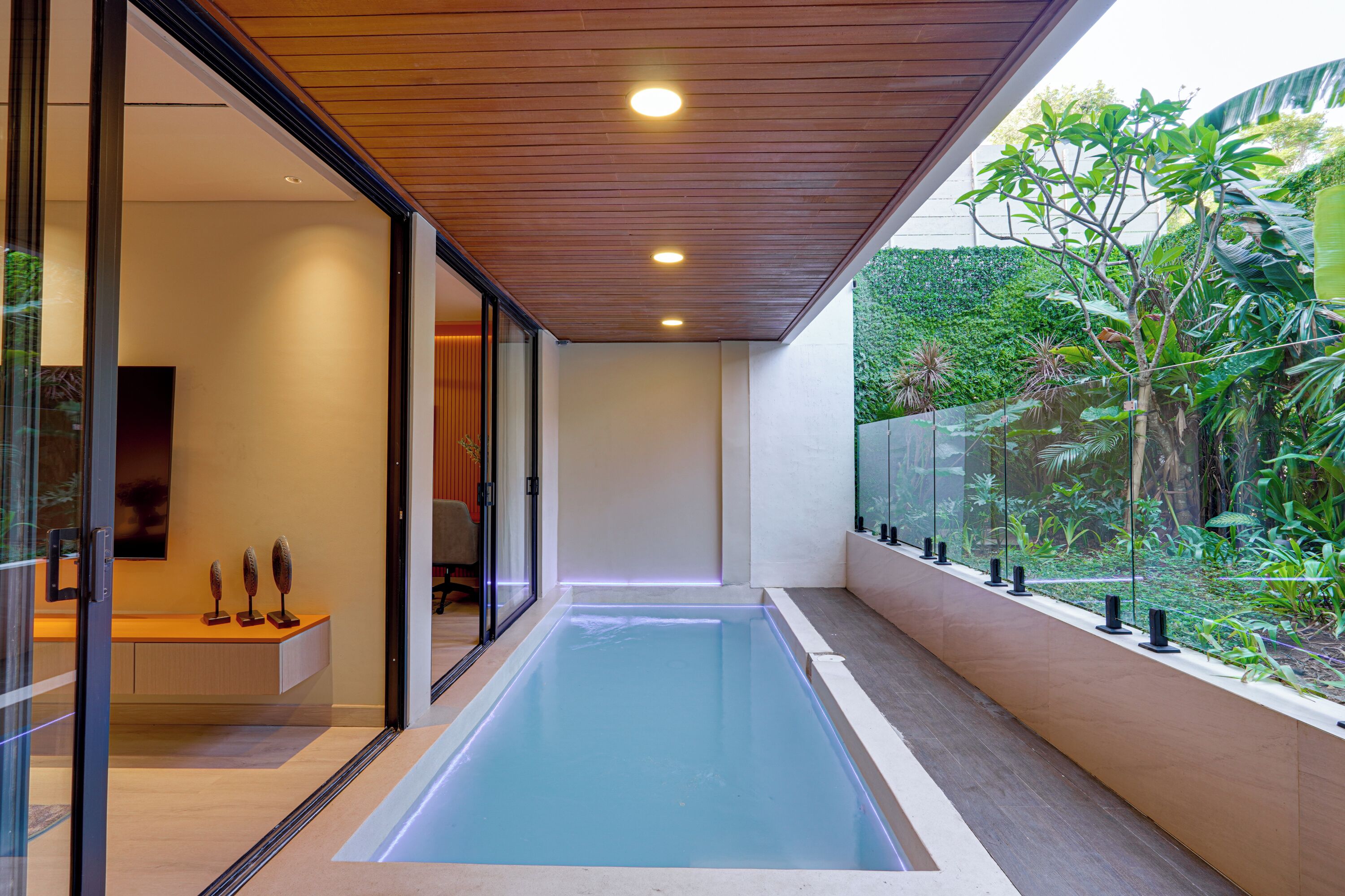 Ein moderner, überdachter Pool mit Holzboden, Glaswänden und Blick auf üppiges Grün.