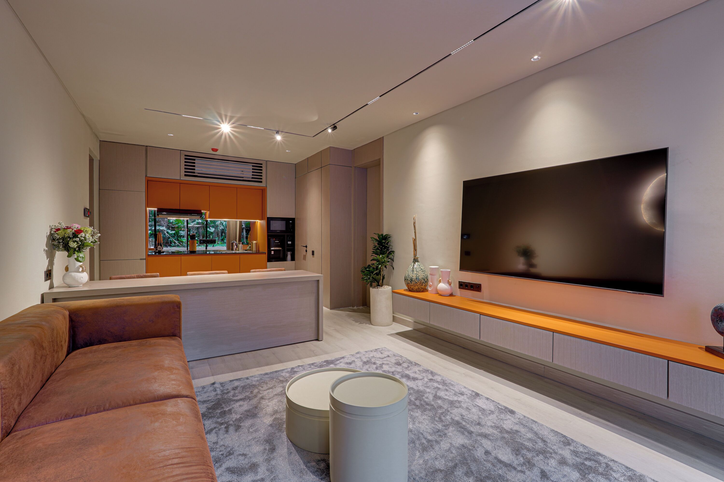 A modern living room with a large flat-screen TV mounted on the wall, a comfortable sofa, and a kitchen area in the background.