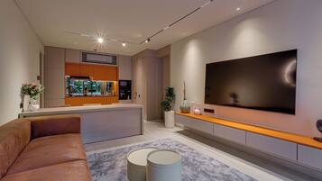 A modern living room with a large flat-screen TV mounted on the wall, a comfortable sofa, and a kitchen area in the background.