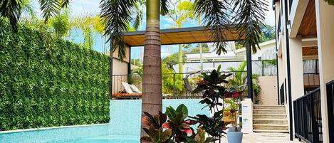 A pool area with steps leading to a covered seating area, surrounded by greenery and a clear blue sky.