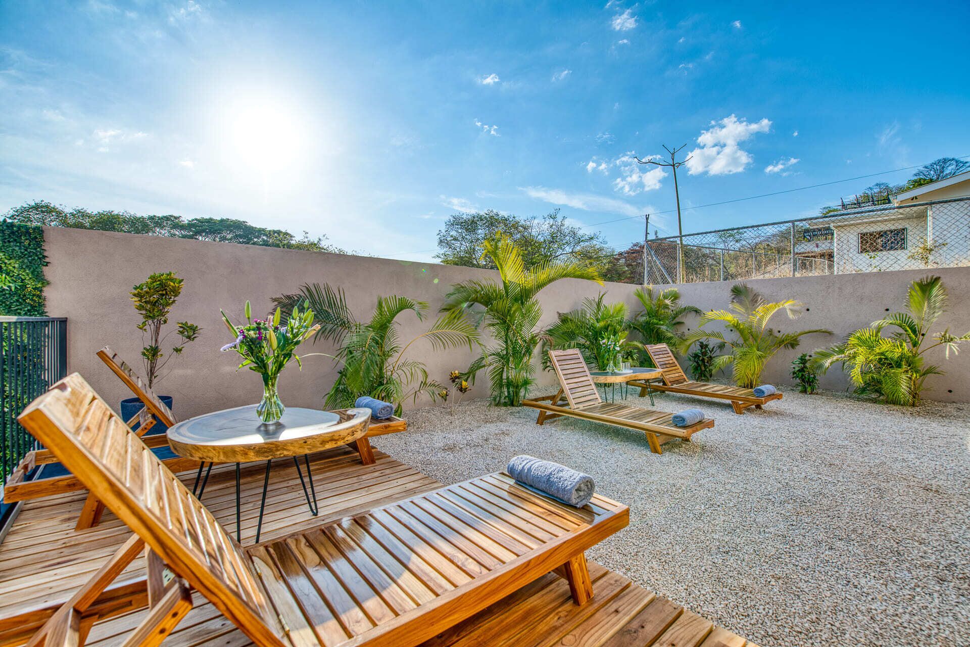 An outdoor patio with wooden lounge chairs and a small round table, surrounded by gravel and various tropical plants.