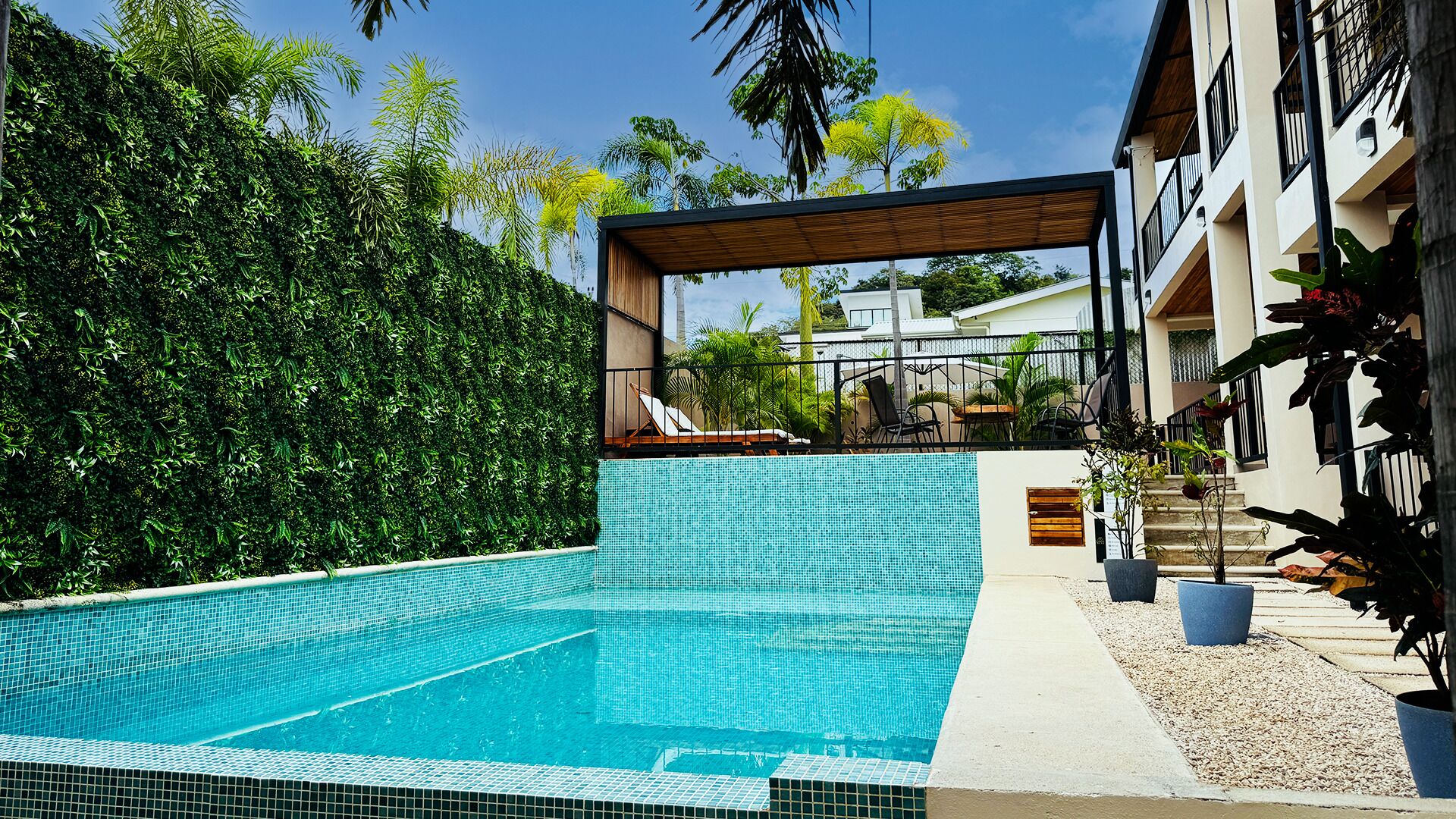 A swimming pool with a tiled deck, surrounded by lush greenery and a covered seating area with lounge chairs.