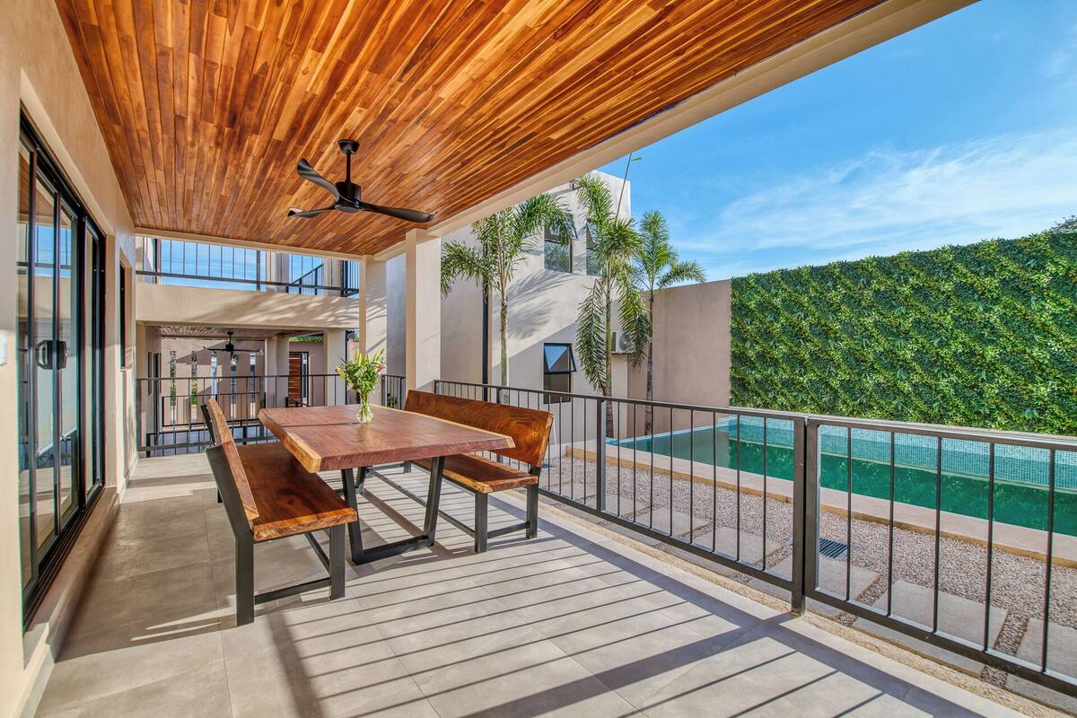 Eine Holzterrasse mit Tisch und Bänken, mit Blick auf einen Swimmingpool und einen üppigen, grünen Heckenstreifen.