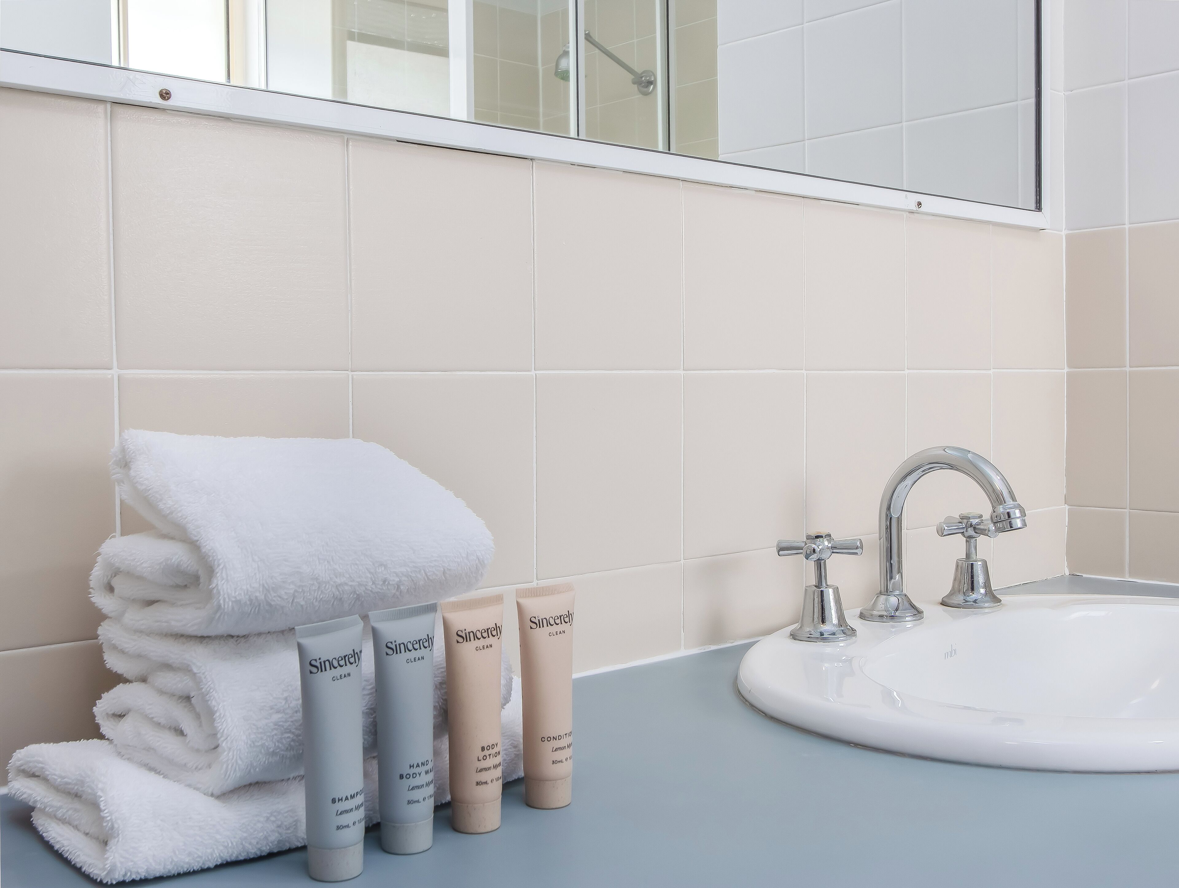 A bathroom with a sink, mirror, and toiletries including a towel and hand lotion.