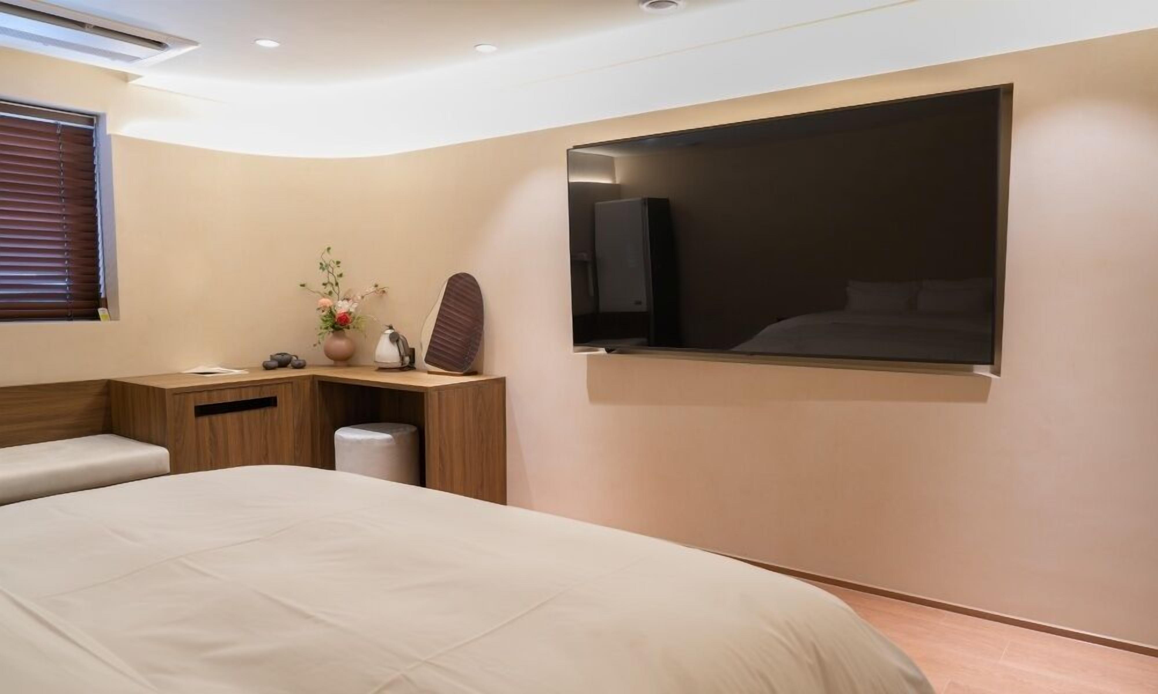 A modern bedroom with a large bed, a desk with a chair, a small trash bin, and a wall-mounted TV.