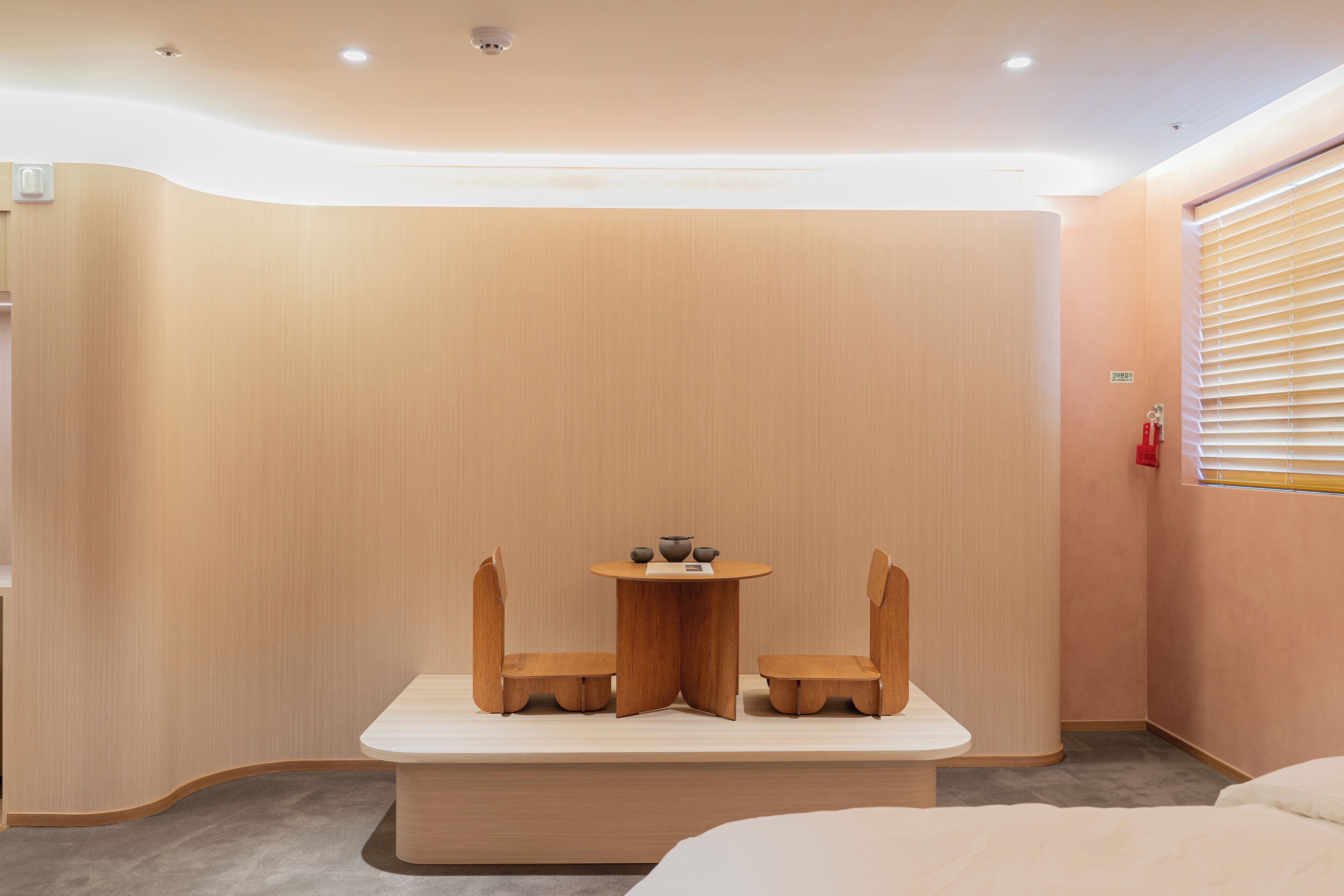 Quarto de hotel moderno com parede de destaque em madeira, uma pequena mesa redonda com duas cadeiras e uma cama em primeiro plano.