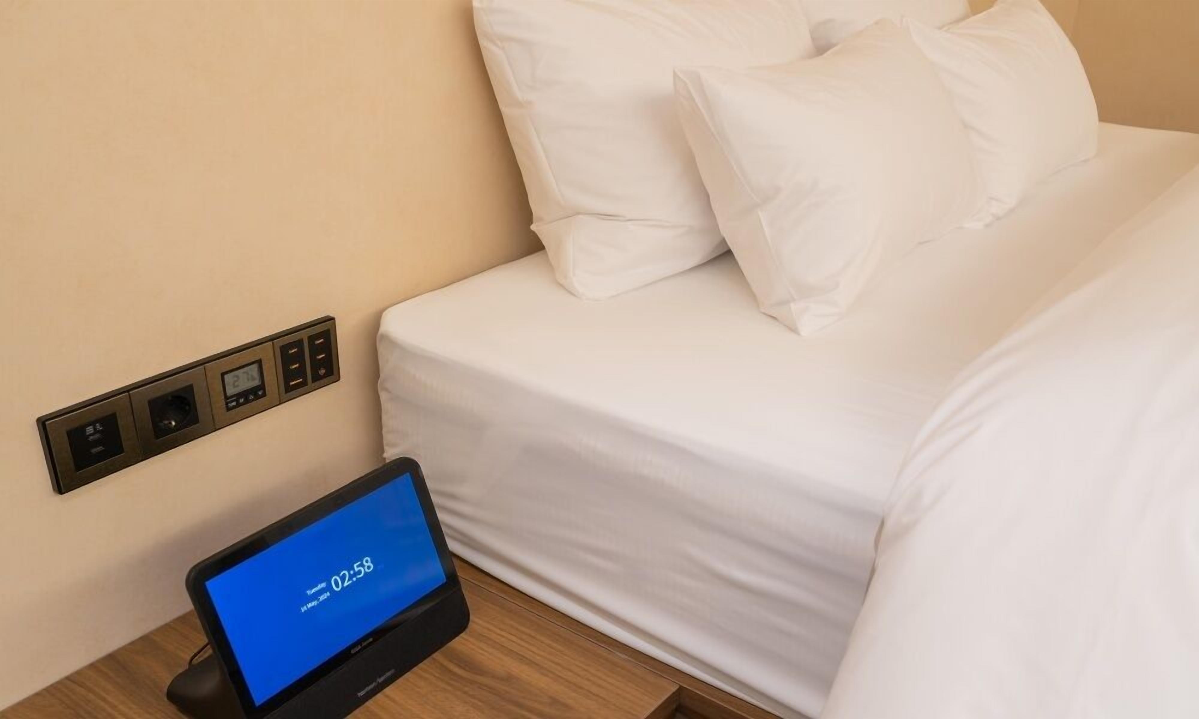 A hotel room with a bed, white bedding, a digital clock, and a control panel on the wall.