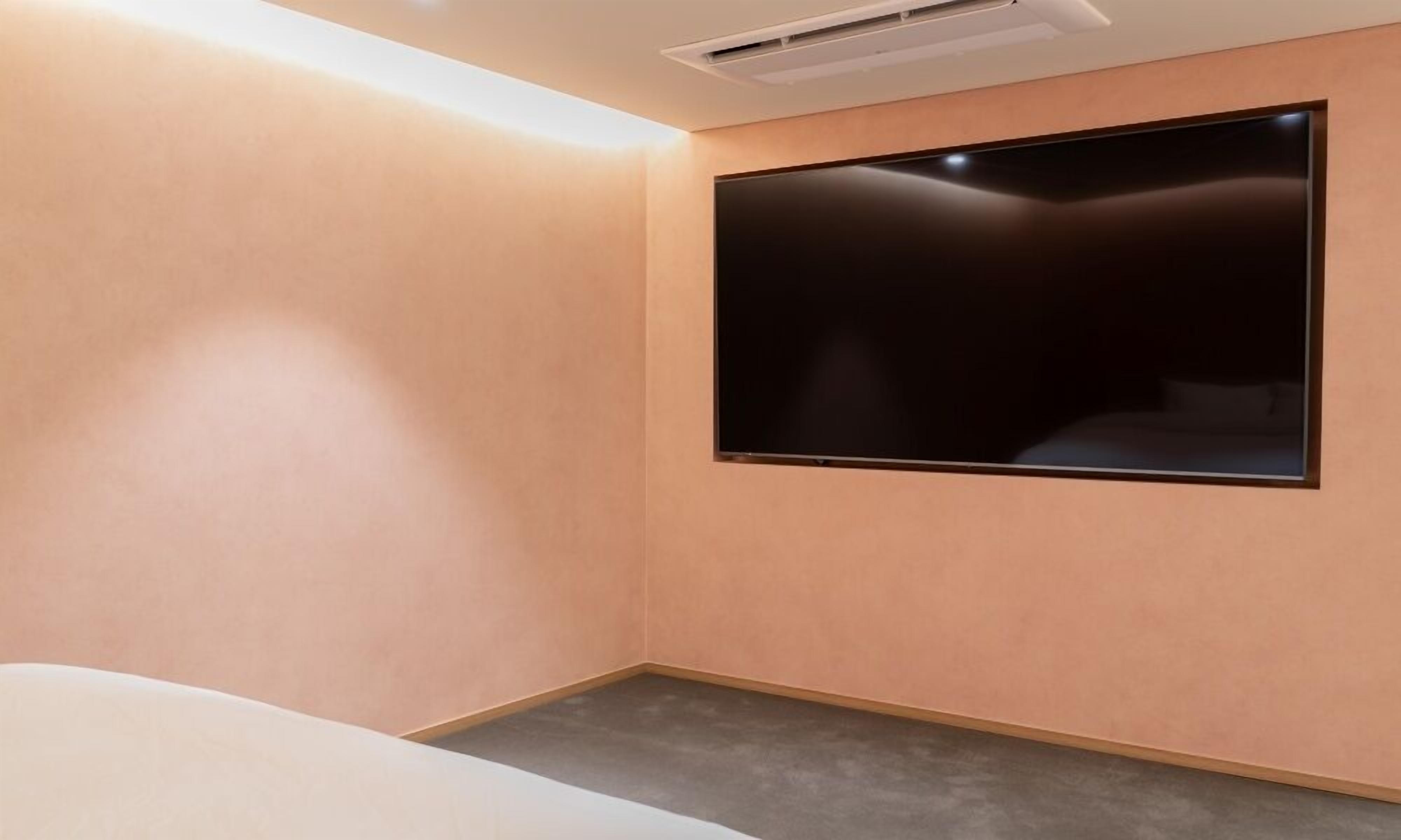 A room with a large flat-screen TV mounted on the wall, a bed with a grey headboard, and a ceiling air vent.