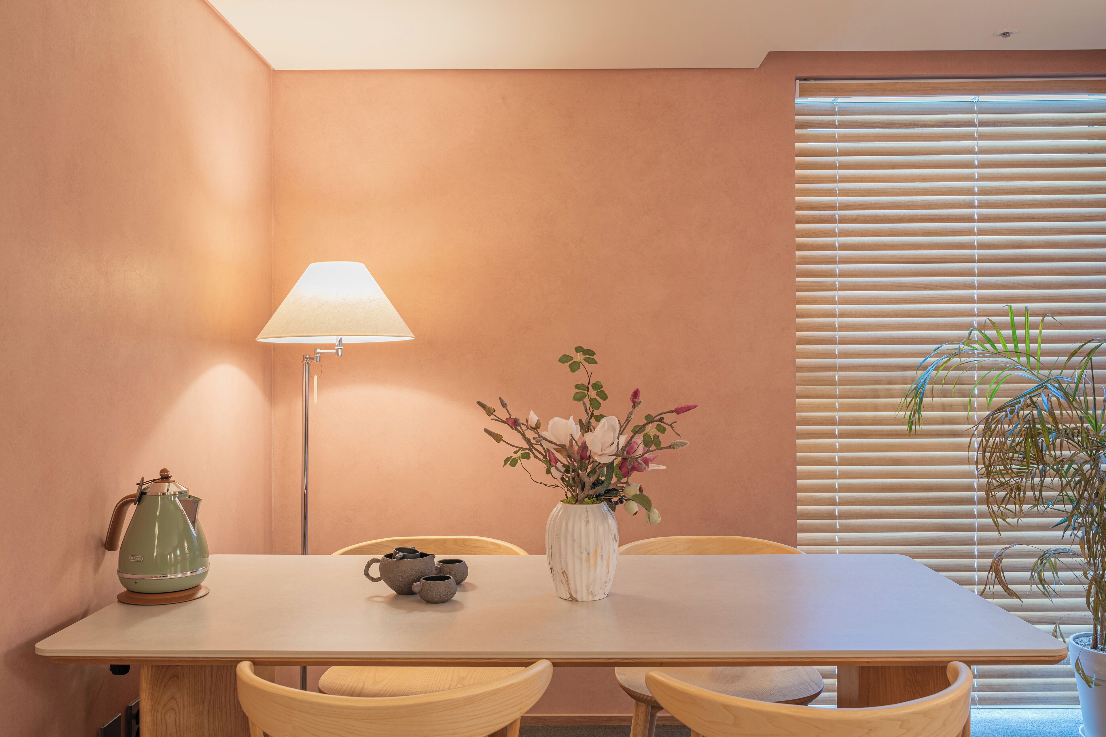 A dining area with a table, chairs, a lamp, a vase with flowers, and a window with blinds.