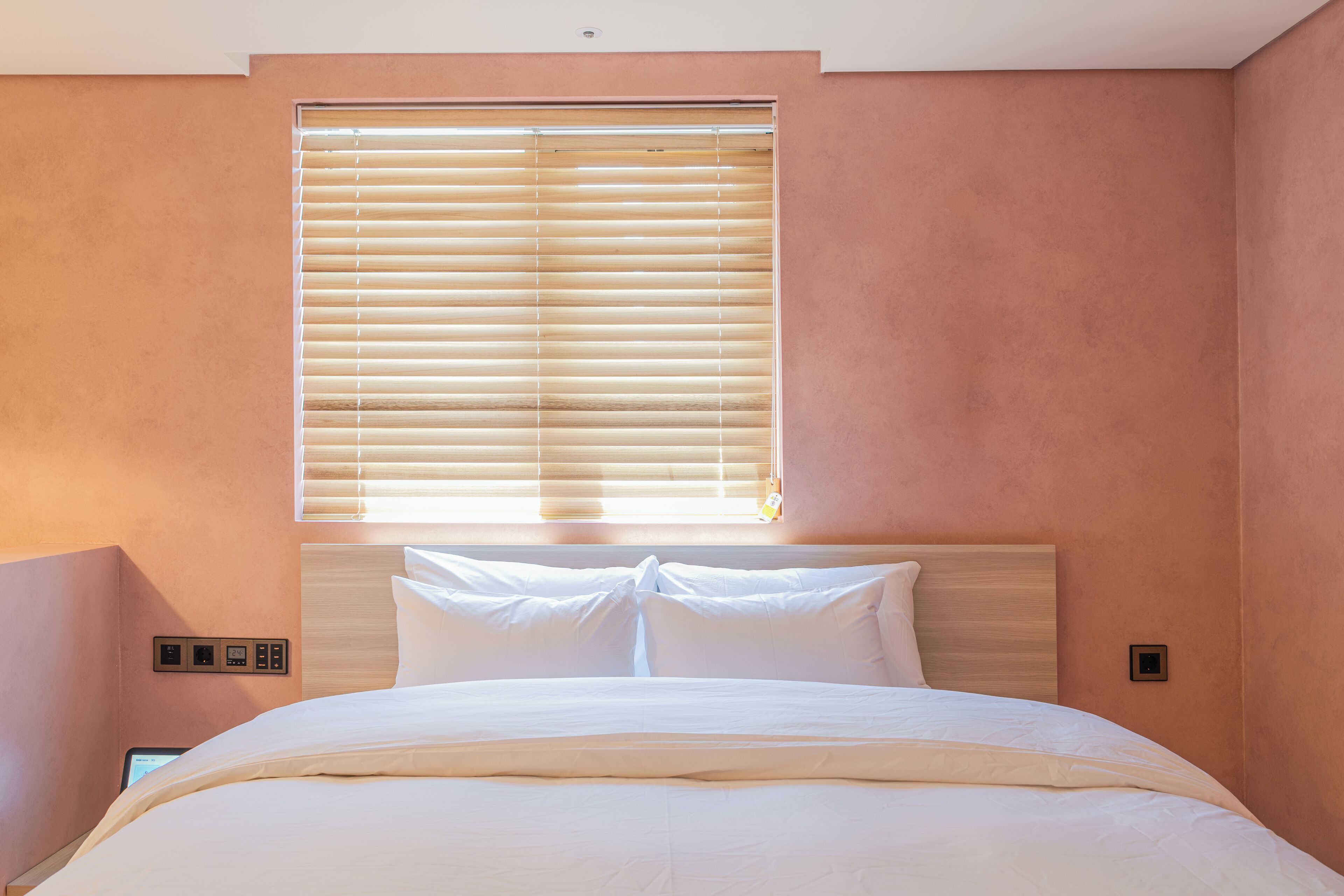 A bedroom with a bed, a window with blinds, and a wall-mounted control panel.