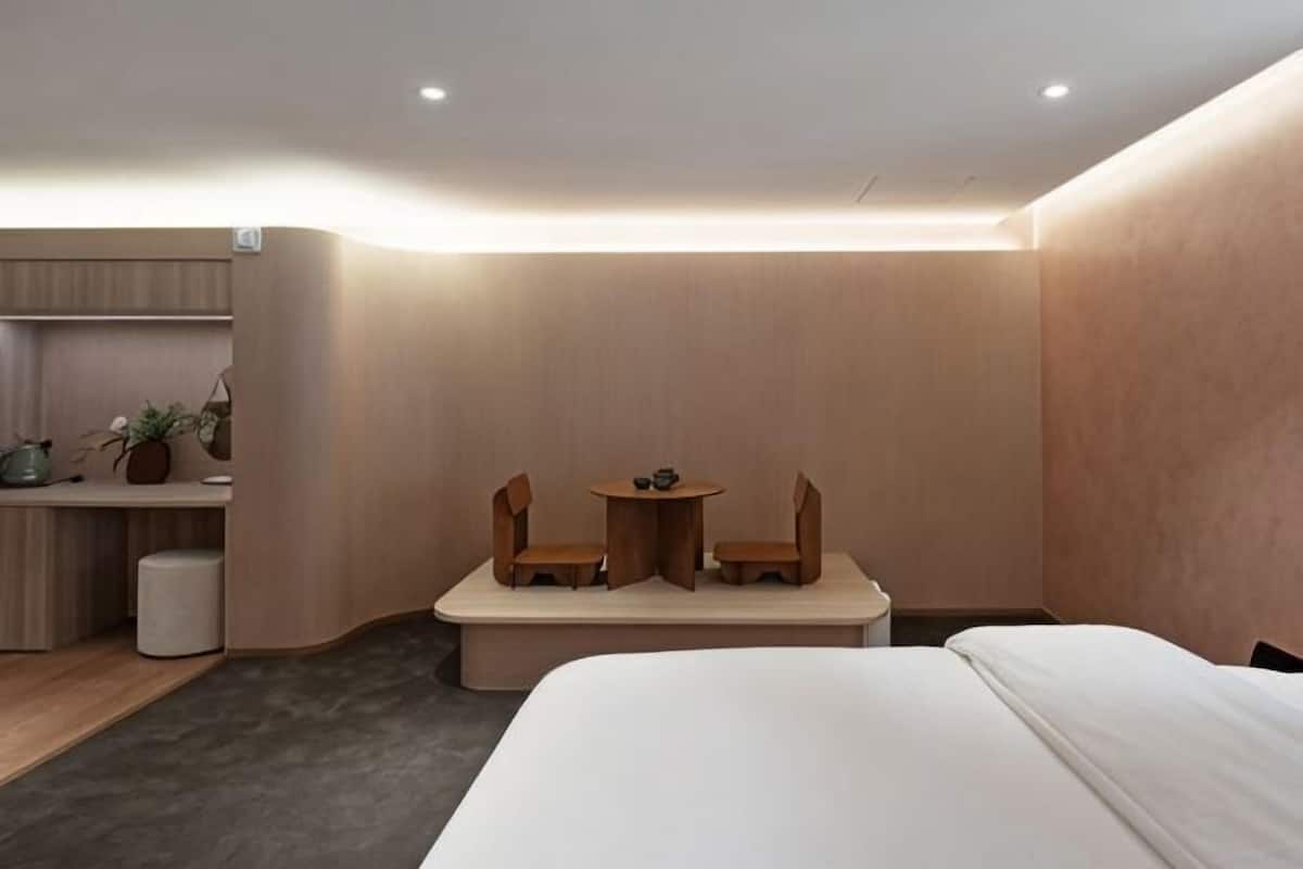 A hotel room with a bed, a small desk with a chair, a table with two chairs, and a plant.