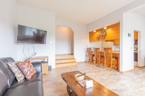 Uma sala de estar moderna com uma TV de tela plana, um sofá de couro e uma mesa de centro de madeira. Uma área de cozinha com armários de madeira e um conjunto de jantar é visível ao fundo.