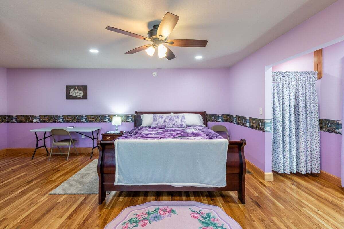 Un dormitorio con cama, escritorio, silla, ventilador de techo y ventana con cortinas.