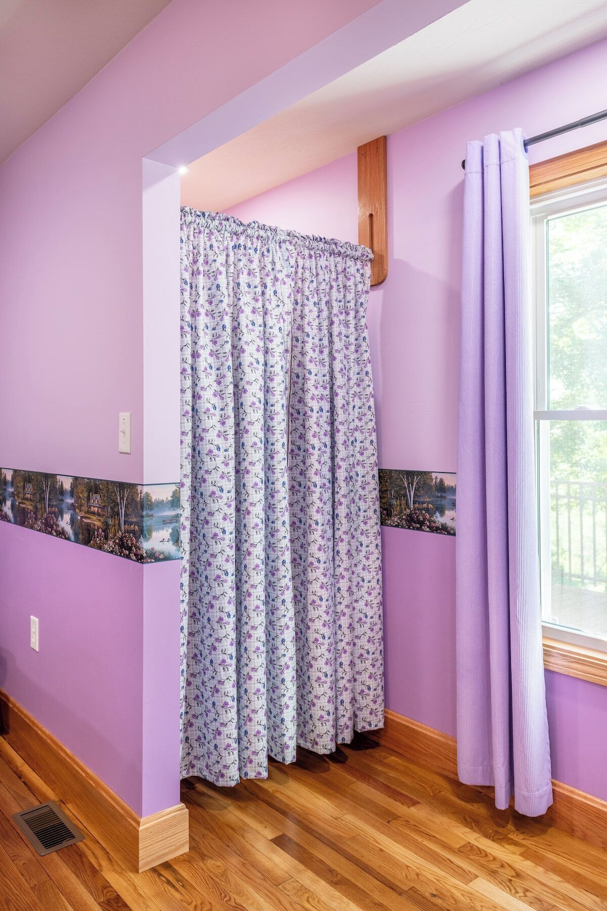 A bathroom with purple walls, a wooden floor, a shower curtain with a floral pattern, and a window with a view of trees.