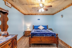 Une chambre avec un plafond en bois, un lit recouvert d’une couverture bleue, une table de chevet avec une lampe et une commode.