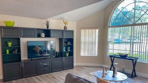 A modern living room with a grey sofa, a TV on a black cabinet, and a large window with blinds. - Luxury Condo Apartment Near Disney Universal 12 (Kissimmee)
