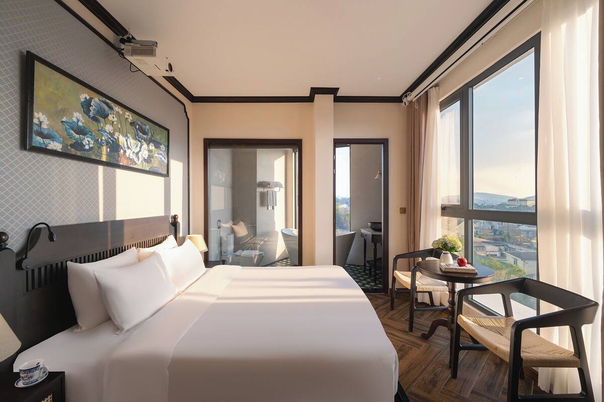 A hotel room with a large bed, a desk, a chair, and a view of the city.