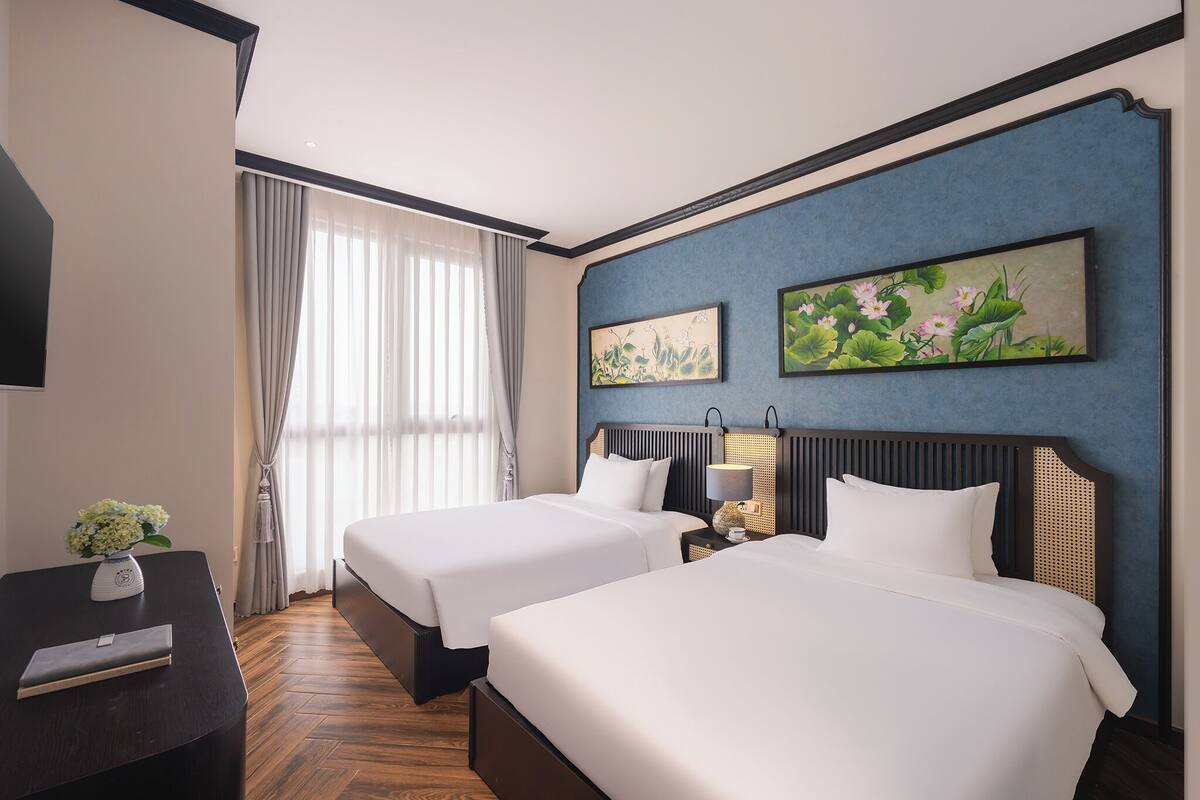 A hotel room with two beds, a desk, a lamp, and two paintings on the wall.