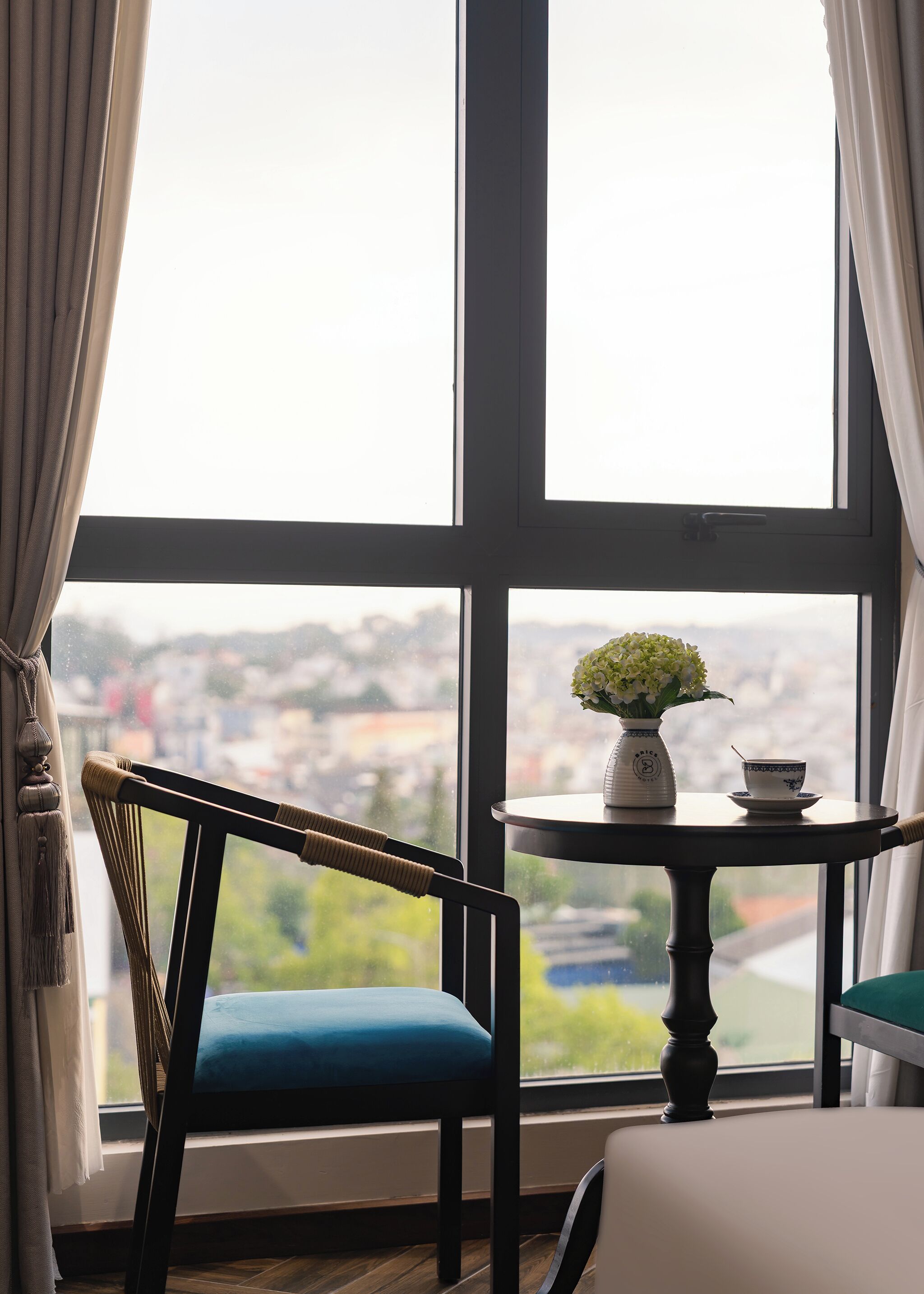 Una habitaci ón con un ventanal grande, una silla, una mesa con un florero y una taza, y vista a un paisaje urbano.