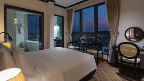 A hotel room with a large bed, a desk with two chairs, a round mirror, and a view of the cityscape through large windows. - BRICS Boutique Dalat (Lam Vien - Da Lat)