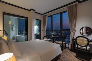 Une chambre d’hôtel avec un grand lit, un bureau avec deux chaises, un miroir rond et une vue sur le paysage urbain grâce à de grandes fenêtres.