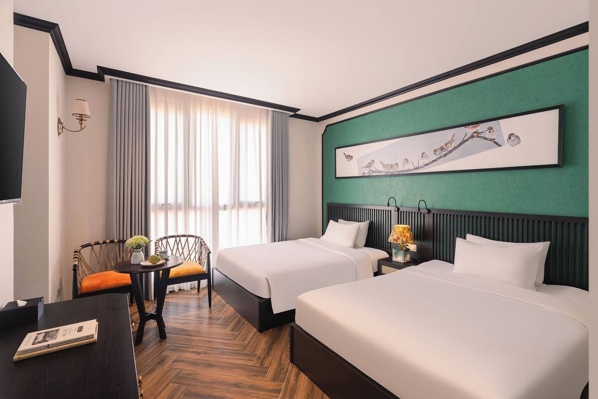 A hotel room with two beds, a desk, a chair, a lamp, a painting, and a window with curtains.