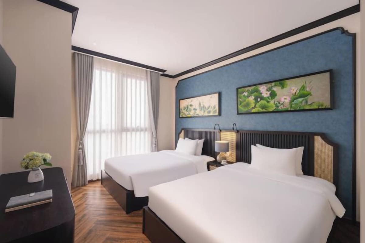 Une chambre d’hôtel avec deux lits, un bureau avec une chaise, une fenêtre avec des rideaux et des œuvres d’art accrochées aux murs.