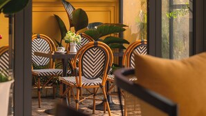A room with a yellow wall, large windows, and a table with chairs. There are plants on the windowsill and a yellow cushion on the table. - BRICS Boutique Dalat (Lam Vien - Da Lat)