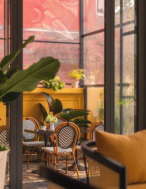 Une pièce avec un mur jaune, de grandes fenêtres et une table entourée de chaises. On peut voir des plantes sur le rebord de la fenêtre et un coussin jaune sur la table.