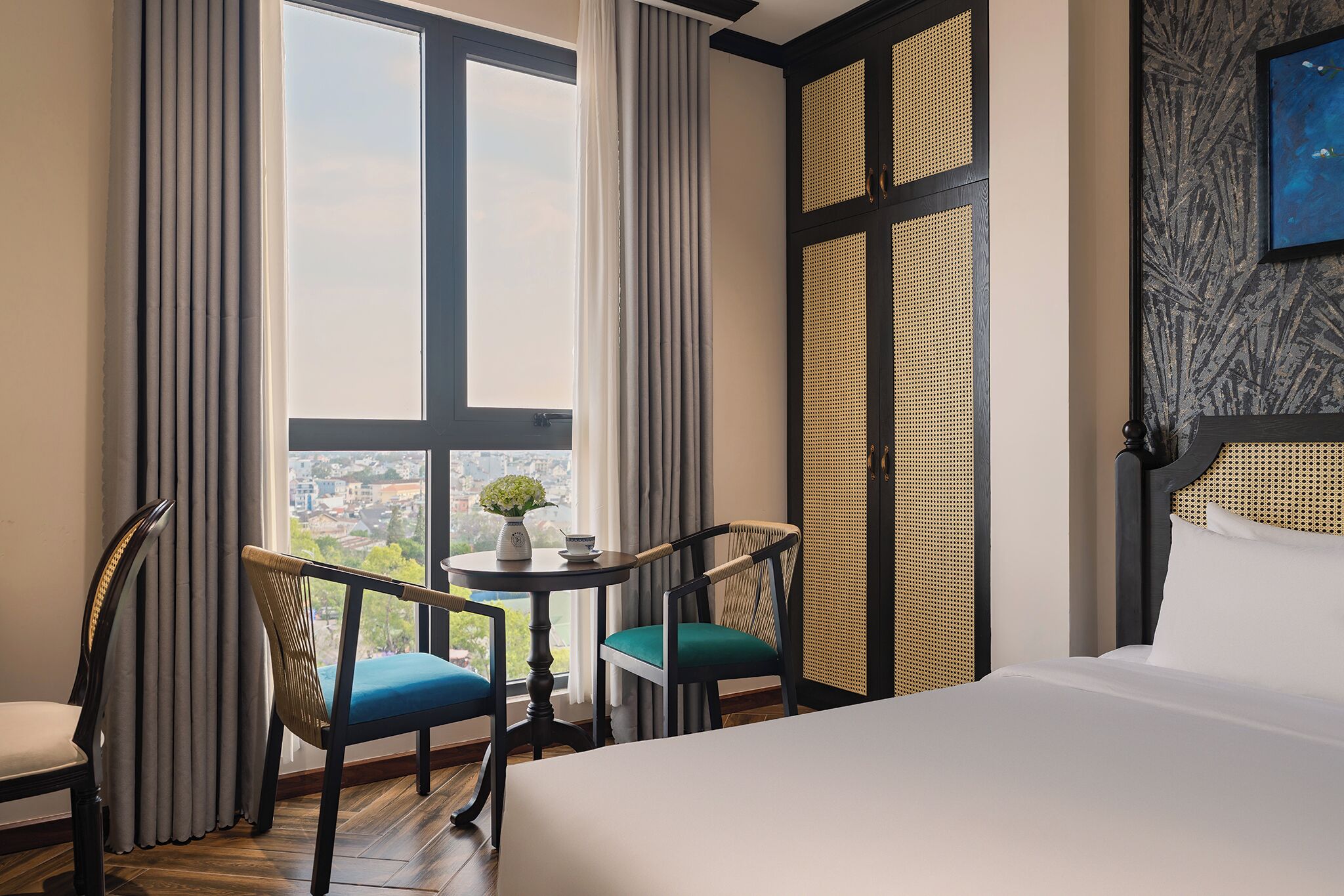 Une chambre d’hôtel avec un lit, deux chaises, une petite table avec un vase et de grandes fenêtres avec des rideaux.